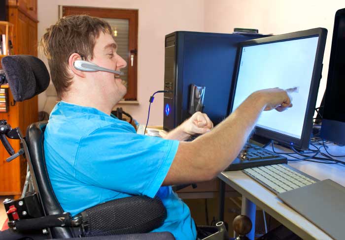 A young man with cerebral palsy sitting in a multifunctional wheelchair using a computer with a wireless headset reaching out to touch the touch screen